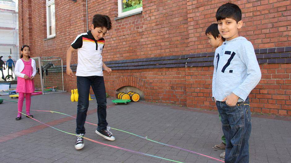  Tarek tanzt den Gummitwist im deutschen Nationaltrikot, während Danah (li.), Astivan (r.) und Demchi auf dem Schulhof der Ferdinand-Heye-Schule zugucken. 
