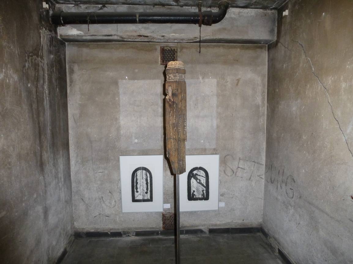  Düster: Der Pranger steht in der Mitte des Raumes. An der Wand Bilder von Menschen am Pranger.  
